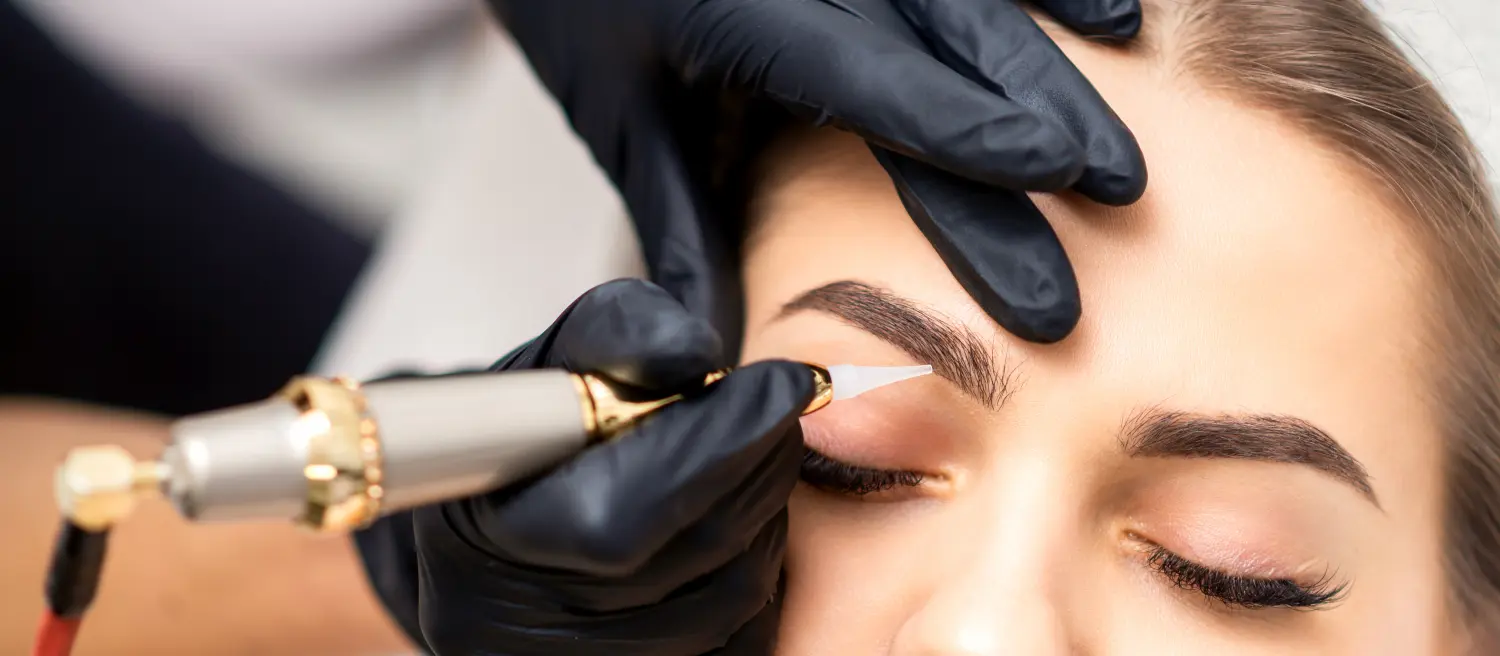 Kosmetikerin trägt mit einer speziellen Tätowiermaschine Permanent Make-up auf die Augenbrauen einer jungen Frau auf